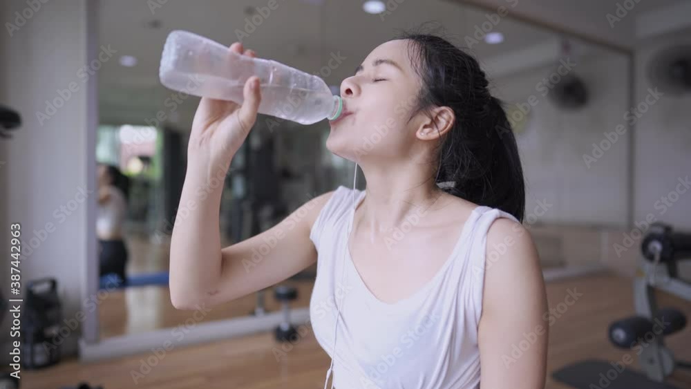 Young pretty asian female drinking water after exercise, refresh feeling, fresh cold water from plastic bottle, thirsty fit woman inside indoor gym, sit down on yoga mat, rehydrated health condition Young pretty asian female drinking water after exercise, refresh feeling, fresh cold water from plastic bottle, thirsty fit woman inside indoor gym, sit down on yoga mat, rehydrated health condition