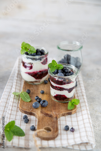 Vasetti di yogurt con yogurt bianco e confettura di mirtilli decorati con mirtilli e foglie di menta su tagliere di legno e mirtilli e menta sparsi sul tavolo