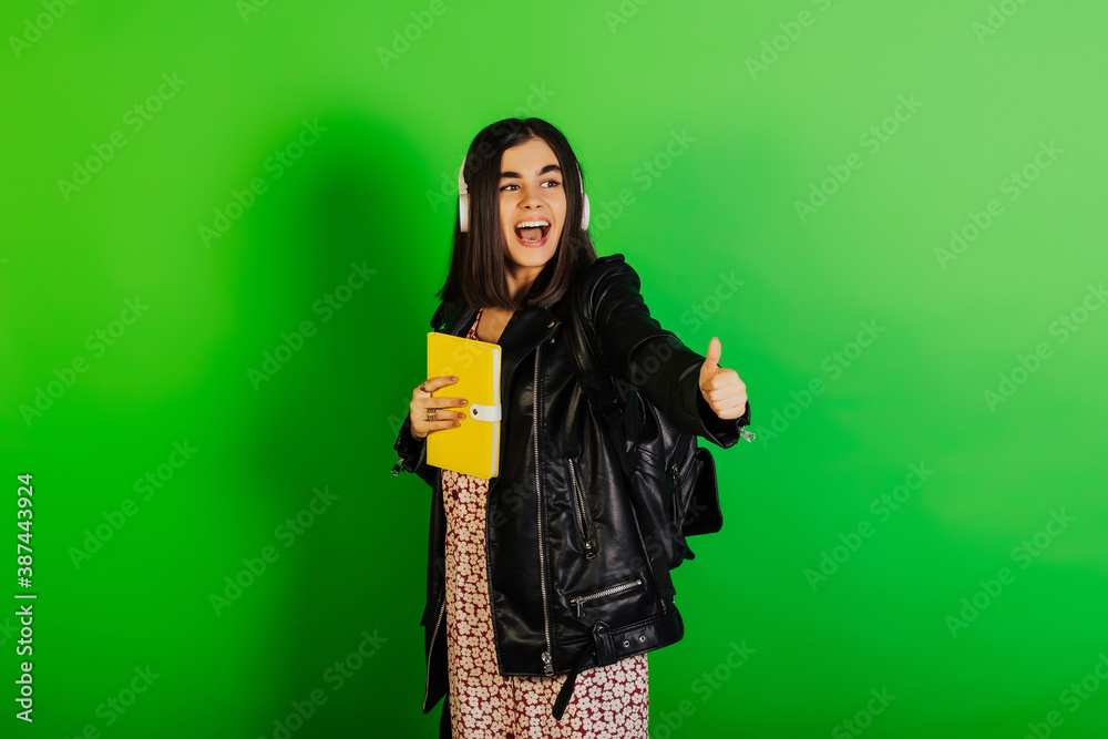 Smiling student girl in black leather jacket with black backpack and headphones hold yellow notebook and shows the sign cool isolated on green background.