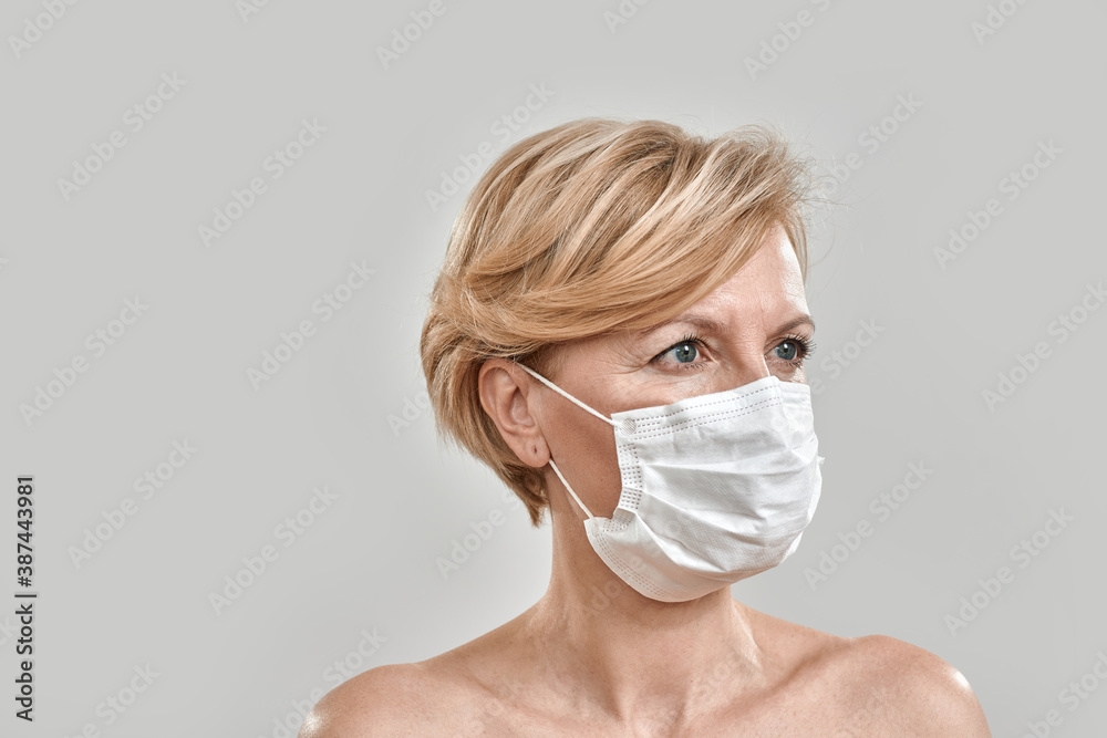 Close up portrait of beautiful middle aged woman wearing white medical mask and looking away isolated against grey background. Pandemic, safety, prevention concept Close up portrait of beautiful middle aged woman wearing white medical mask and looking away isolated against grey background. Pandemic, safety, prevention concept