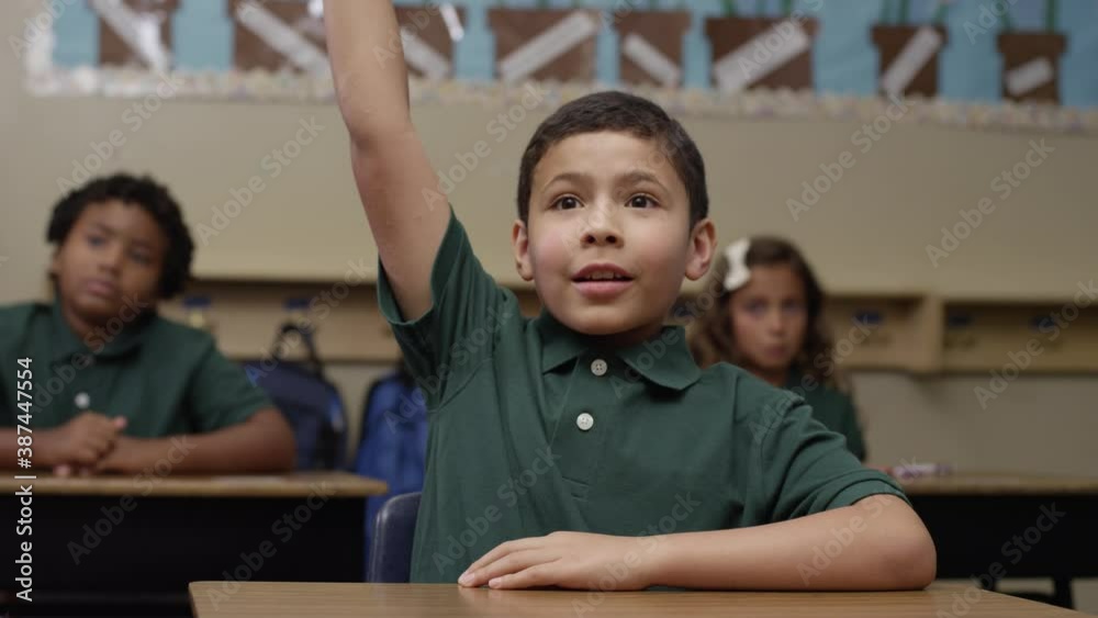 Primary students in uniforms sit in class as happy brown boy raises his hand