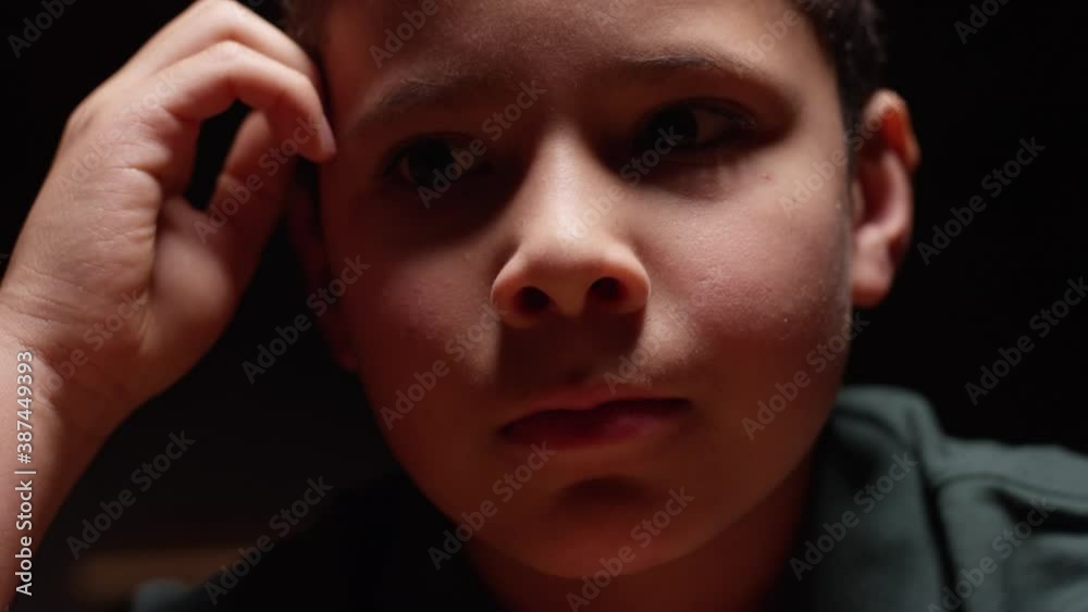 Young hispanic boy sitting at desk in the dark with a sad, lonely expression Young hispanic boy sitting at desk in the dark with a sad, lonely expression