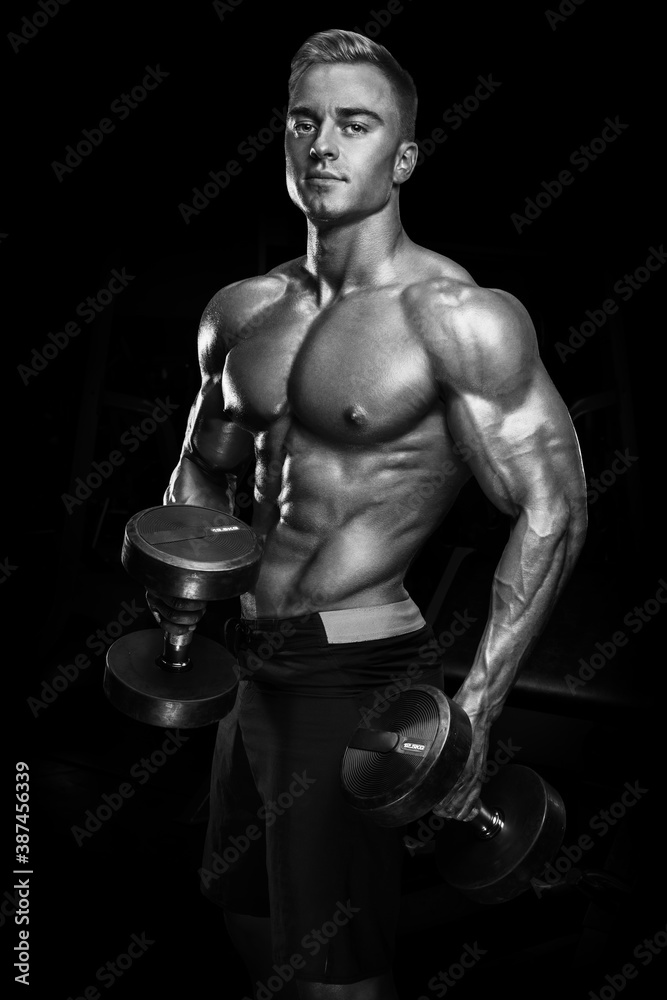 Perfect fit athletic guy workout with dumbbells. Handsome power athletic man in training pumping up muscles with dumbbells in a gym. Fitness muscular body isolated on dark background. Perfect fit athletic guy workout with dumbbells. Handsome power athletic man in training pumping up muscles with dumbbells in a gym. Fitness muscular body isolated on dark background.