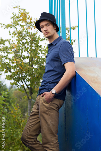 a young, attractive guy in a hat, blue shirt, and beige trousers is leaning against a building
