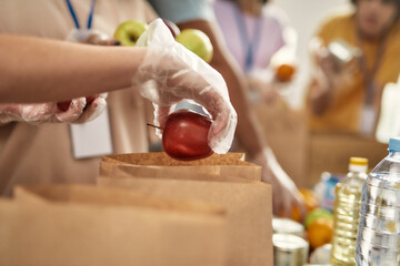 Close up of hand of volunteer in glove holding an apple while collecting, sorting food for needy people in paper bags, Team working together on donation project