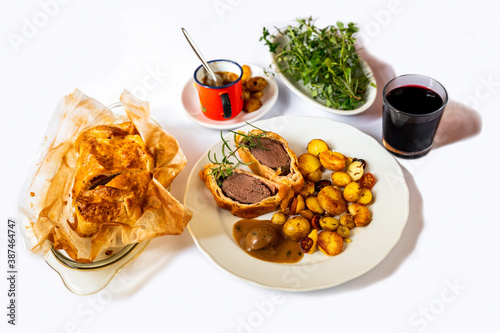 Baked deeer Wellington, portion of meal on plate, cutlery and red wine,pot with sauce and herb. photo