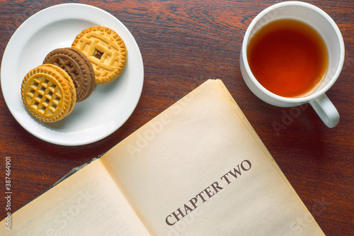 Cup of tea, cookies on a saucer and an open book on a wooden table. Reading during breakfast.