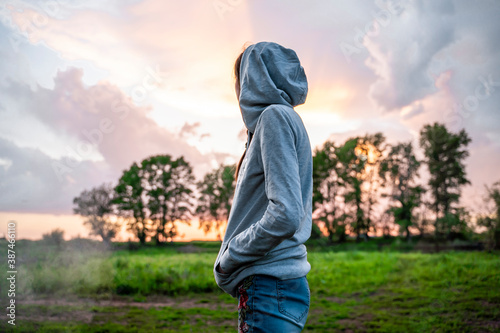 A young woman in a hood stands sideways and looks at the sunset. Forest