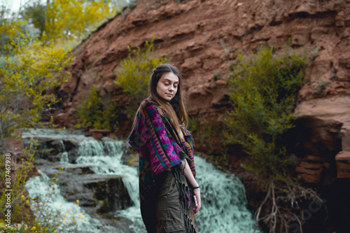 Beautiful young woman in a poncho stands in front of a waterfall with closed eyes
