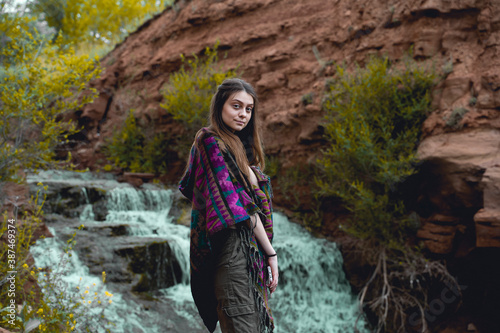 A beautiful young woman in a poncho stands in front of a waterfall