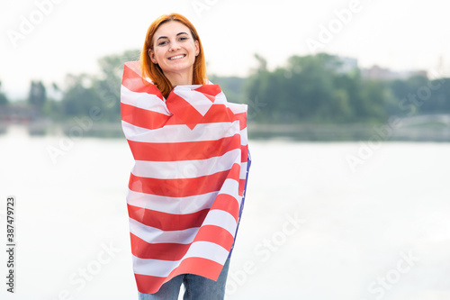 Portrait of happy red haired girl with USA national flag on her shoulders. Positive young woman celebrating United States independence day. International day of democracy concept.
