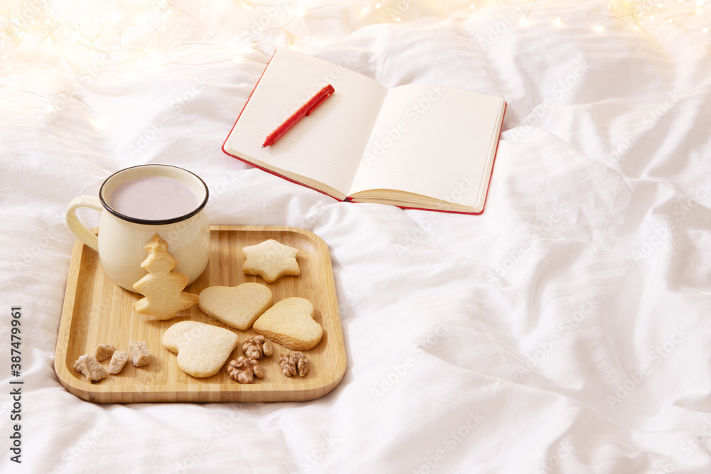 Concept of lazy time at home, holidays. Future planning female theme. Tray with Christmas cookies and cocoa on the bed. Nearby is an open notebook. cotton white blanket, bokeh lights on background