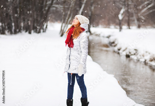 Outdoors fashion lifestyle portrait of charming young woman enjoying snowfall in snowy forest. Wearing silver down jacket, red scarf and mittens. Winter river landscape. Enjoying nature and snowfall