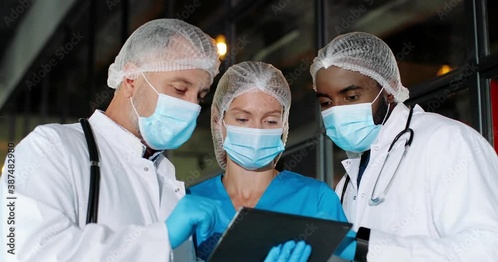 Mixed-races males and female doctors in hats, medical masks and gloves standing in hospital, talking, using tablet device. Multi ethnic men and woman, medics discussing diagnosis covid-19. Coronavirus Mixed-races males and female doctors in hats, medical masks and gloves standing in hospital, talking, using tablet device. Multi ethnic men and woman, medics discussing diagnosis covid-19. Coronavirus