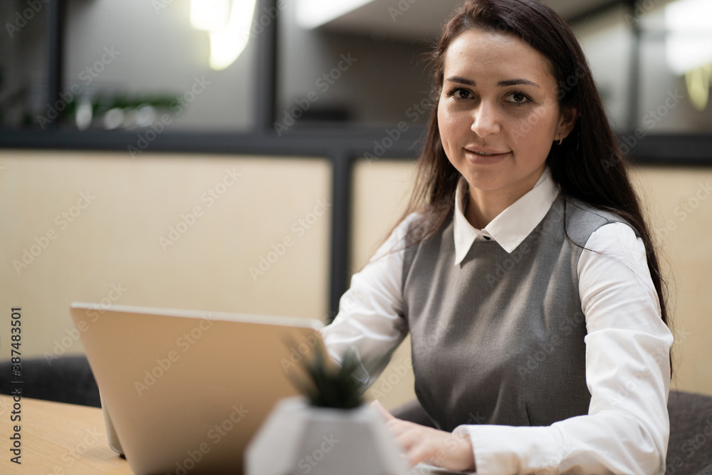 European-looking brunette sitting in a coworking office, looking at the camera. confident woman blogger working on the laptop writing new posts for your social networks and to their subscribers.