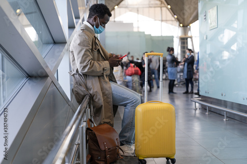 Afro-American traveler man in trench coat wear face protective mask, sitting in airport terminal or railway station, using mobile phone, waiting for flight and boarding. New normal, covid-19 pandemic.