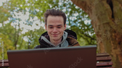 Serious guy young man. student works on a laptop in the park. Remote work and study concepts. Close-up shot of a young programmer rejoicing at the camera. The concept of a successful teenager in the