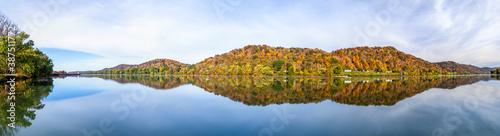 Hills covered in colorful fall foliage are reflected on the still water of the beautiful Ohio River. Monroe County, Ohio is viewed from the river bank at Paden City, West Virginia.