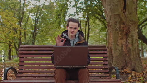 Medium shot of handsome young man working on laptop computer sitting outdoors in park, then looks at the camera and shows OK.Work alone is far from the office,freelancer