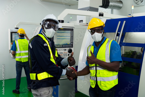 technician supervisor and factory technicians shake hand in factory with protective safety mask, glasses, helmet and a sign of social distance during coronavirus or covid 19 outbreak