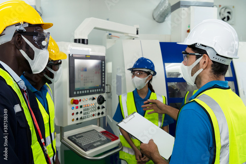 Male technician supervisor discuss with a group of factory technicians in factory with protective safety mask, glasses and helmet as new normal lifestyle during coronavirus or covid 19 outbreak