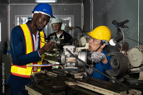 Male technician inspector discusses with male factory mechanic expert in front of a repair machine in factory maintenance office with protective glasses, gloves and helmet in factory uniform