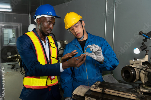 Male technician inspector discusses with male factory mechanic expert in front of a repair machine in factory maintenance office with protective glasses, gloves and helmet in factory uniform