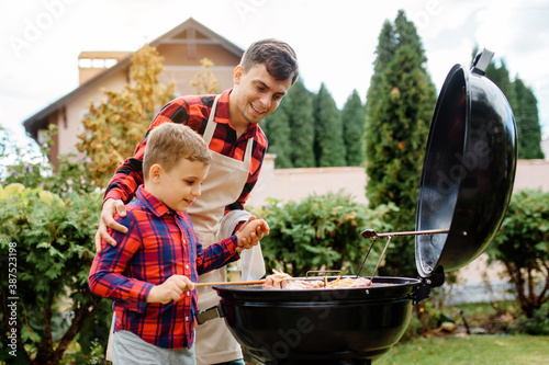 Young father and his son are cooking barbecue on an outdoor grill in their home backyard and having fun.