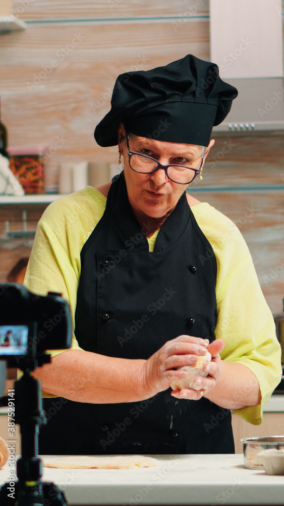 Fototapeta premium Grandmother making cooking vlog, recording herself on camera. Retired blogger chef influencer using internet technology communicating, shooting blogging on social media with digital equipment