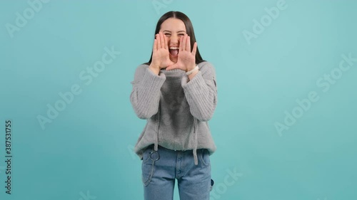 Young woman that is shouting angry out loud with hands over mouth, isolated over turquoise background.