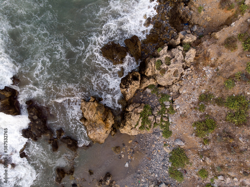 Obraz premium Aerial top down photo of foaming waves crashing against rocky shore, big stones on coastline. Aegean sea, nature of Crete, Greece. Sea natural patterns, background wallpaper.