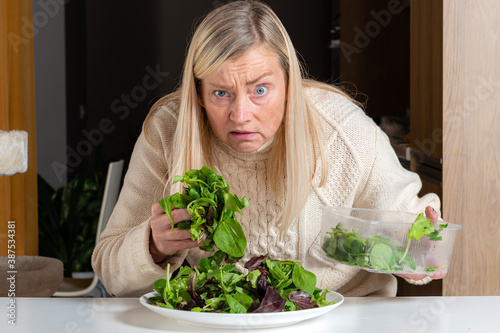 middle aged blonde woman with dissatisfied facial expression preparing green salad in the kitchen