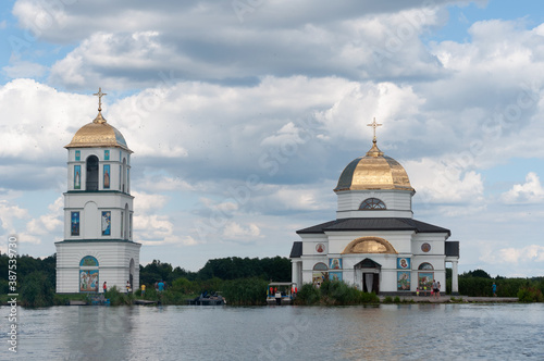 beautiful view of submerged Orthodox Church surrounded by water, trees and reeds under blue sky with clouds