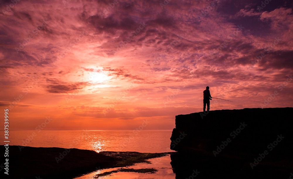 Shore fishing man silhouette on a flat rock at sunset with clouds and purple orange sky near the black sea turkey