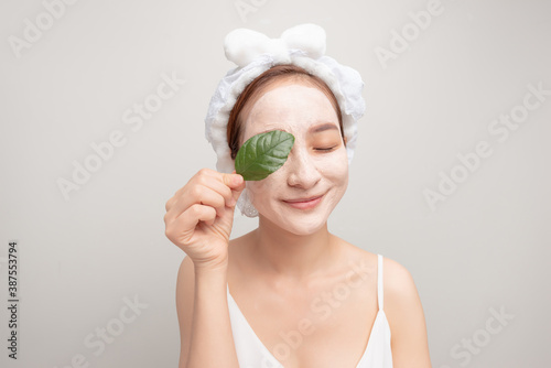 Beauty portrait of woman in towel on head with white nourishing mask or creme on face and green leaf in hand, photo