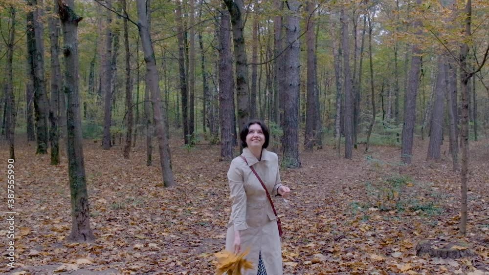 A middle-aged woman, a brunette in a light light raincoat, with a handbag over her shoulder, throws an armful of yellow leaves up and rejoices. slow motion. Park autumn day A middle-aged woman, a brunette in a light light raincoat, with a handbag over her shoulder, throws an armful of yellow leaves up and rejoices. slow motion. Park autumn day