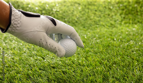 Hand in golf glove holding a golfball, green course lawn background, close up view.