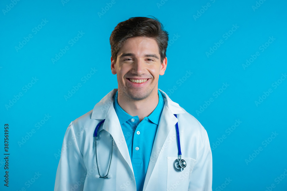 Handsome doctor man in medical wear smiling on blue studio background. Cheerful guy laughing and looking to camera.