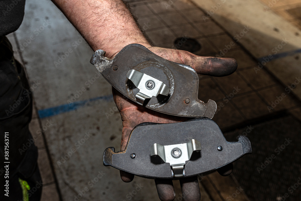 A car mechanic holds in a dirty, open hand a new and old brake pad, visible rear part of the pads.