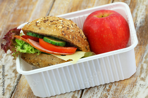 Healthy school lunchbox containing brown roll with smoked sausage with lettuce, tomato and cucumber and red apple
