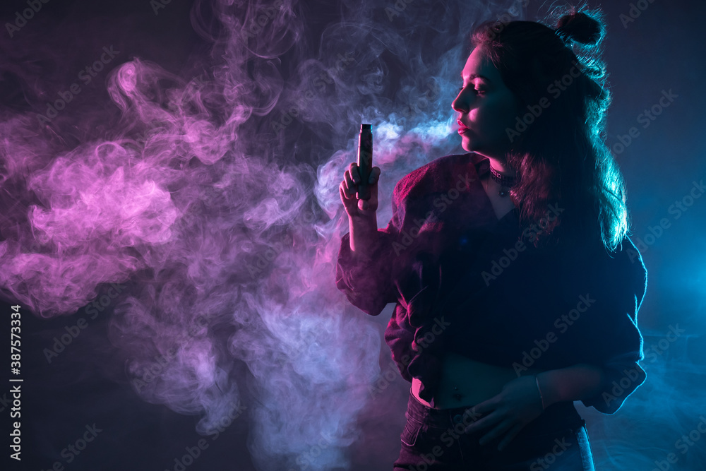 Woman exhales smoke from vape. Vaper girl in dark. Neon light illuminates a vaper woman. Vaping concept. Puffs of smoke from vape next to girl. E-Cigarette as a vaping symbol. Vape device in her hand