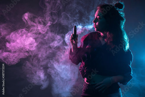 Woman exhales smoke from vape. Vaper girl in dark. Neon light illuminates a vaper woman. Vaping concept. Puffs of smoke from vape next to girl. E-Cigarette as a vaping symbol. Vape device in her hand