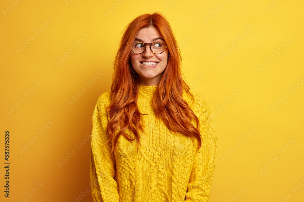 Positive good looking young woman has natural ginger hair looks with happy mysterious expression aside bites lips ponders over nice offer dressed in casual jumper isolated on yellow background Positive good looking young woman has natural ginger hair looks with happy mysterious expression aside bites lips ponders over nice offer dressed in casual jumper isolated on yellow background