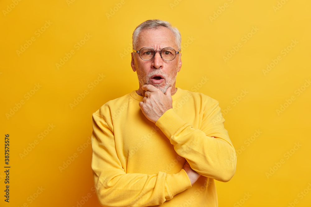 Shocked surprised bearded European man hears unexepcted news holds chin and keeps mouth opened looks with astonishment at camera wears glasses casual sweater isolated over yellow background. Shocked surprised bearded European man hears unexepcted news holds chin and keeps mouth opened looks with astonishment at camera wears glasses casual sweater isolated over yellow background.