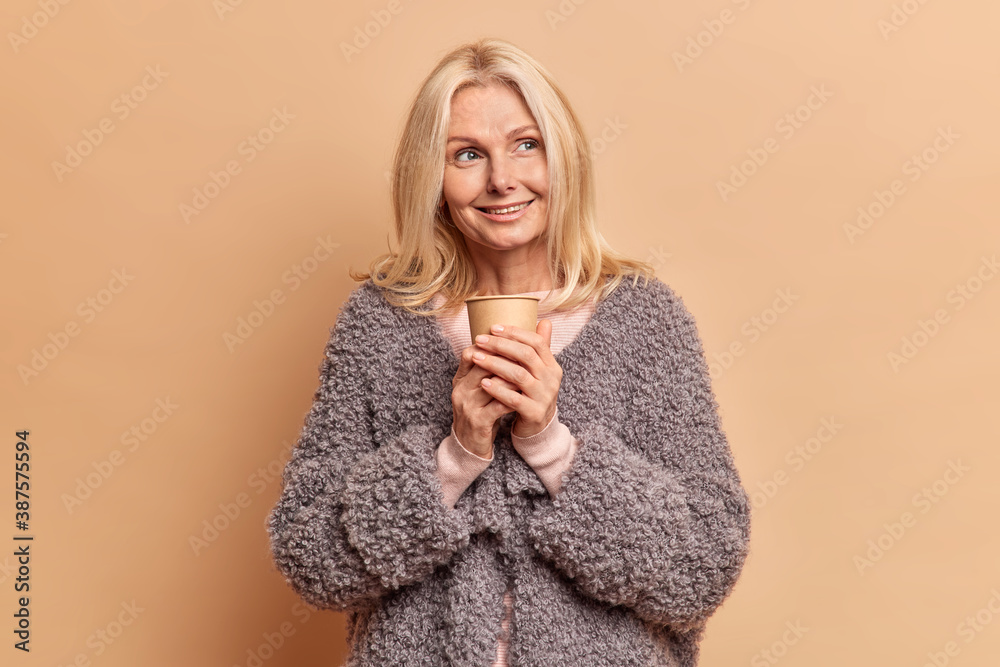 Beautiful tender woman has pleased thoughtful expression looks away dreams about rest poses with paper cup of coffee enjoys drinking aromatic beverage stands against beige studio background. Beautiful tender woman has pleased thoughtful expression looks away dreams about rest poses with paper cup of coffee enjoys drinking aromatic beverage stands against beige studio background.