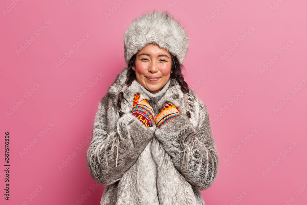 Isolated shot of happy woman smiles positively wears warm fur winter coat and hat has two combed pigtails models against rosy background. Pleased beautiful resident of tundra or far north Isolated shot of happy woman smiles positively wears warm fur winter coat and hat has two combed pigtails models against rosy background. Pleased beautiful resident of tundra or far north
