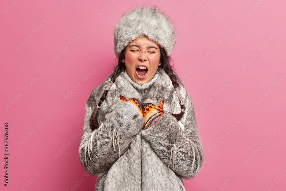 Indigenous circumpolar woman yawns and has sleepy expression wears winter clothes lives at greenland poses against rosy background. Tired Nomadic nenet female dressed in grey fur coat and hat Indigenous circumpolar woman yawns and has sleepy expression wears winter clothes lives at greenland poses against rosy background. Tired Nomadic nenet female dressed in grey fur coat and hat