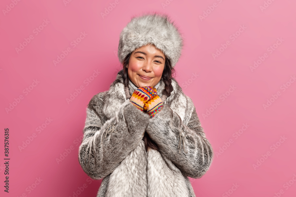 Cute smiling Eskimo woman dressed in winter fur outerwear keeps hands together looks with charming expression poses against rosy background. Tundra inhabitant adapted to severe cold conditions Cute smiling Eskimo woman dressed in winter fur outerwear keeps hands together looks with charming expression poses against rosy background. Tundra inhabitant adapted to severe cold conditions