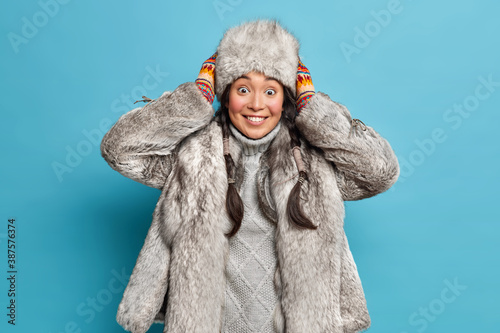 Smiling northern woman of nenets nationality keeps hands on warm fur hat being in good mood has two pigtails isolated over blue background. Indigeous people of North. Lady wears eskimo wear. photo