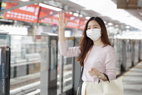 Asian business woman in casual clothes wearing face mask raised her hand to greet friends while waiting for the train to go to work on the platform station. New normal lifestyle in city concept.